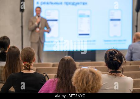Business speaker presents slides to seated audience in modern conference room, attendees listen and take notes, professional seminar atmosphere. Stock Photo