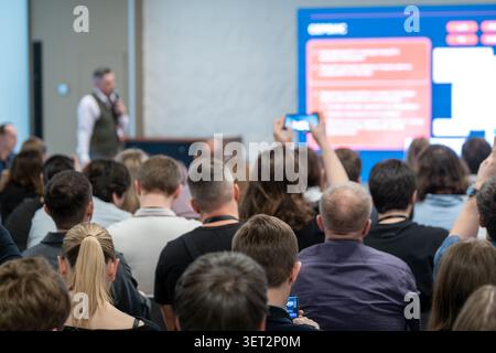 Diverse audience watching business presentation, attendees record speaker on phones, corporate conference setting, focus on engagement and learning. Stock Photo
