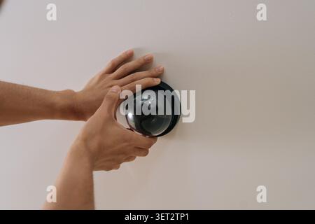 Close-up view of male hands installing black dome surveillance camera on white ceiling. Concept of home security, protection, monitoring, smart home Stock Photo