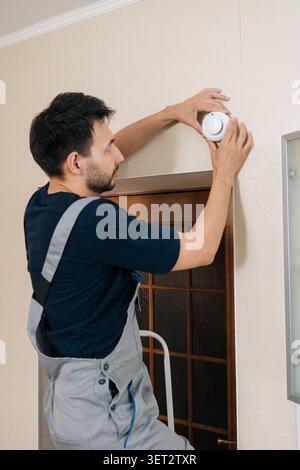 Man in overalls installing smoke detector on interior wall, professional handyman performing home maintenance for fire safety, alarm mounting and Stock Photo