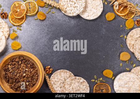 Healthy breakfast ingredients, including granola in a wooden bowl, rice cakes, dried oranges, apricots, pumpkin seeds, and walnuts, arranged on a dark Stock Photo
