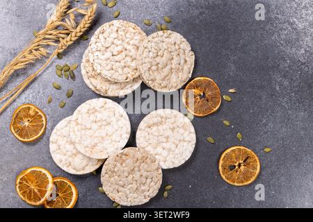 Puffed rice cakes displaying a wholesome snack concept, arranged with oat ears, green pumpkin seeds, and decorative dried orange slices on a dark grey Stock Photo