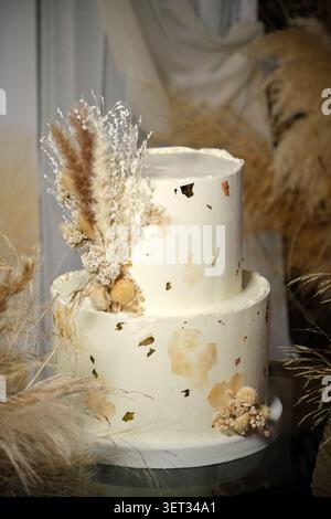 An elegant two-tier wedding cake decorated with gold leaf and dried flowers, displayed in a rustic boho setting with a soft, romantic atmosphere. Stock Photo