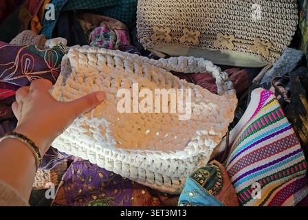 A person holds a white crocheted handmade handbag among other colorful fabric items. Stock Photo