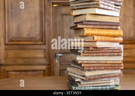 A tall stack of old books sits on a wooden table. The books are worn and have different covers. A wooden wall and door are visible in the background. Stock Photo