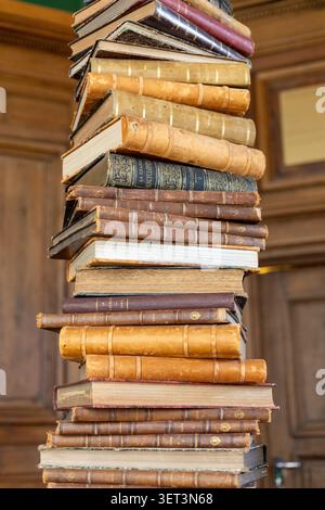 A tall stack of old books stands in a library. The books have worn covers and are stacked on top of each other. Wooden shelves provide a backdrop, and Stock Photo