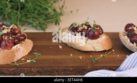 Crispy baguette slices topped with creamy goat cheese, roasted red grapes, crushed walnuts, and fresh thyme sprigs on a rustic wooden board. The compo Stock Photo
