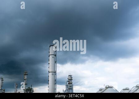 A tall distillation tower and spherical storage tanks at an oil refinery under dramatic dark storm clouds Stock Photo