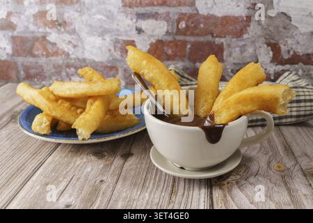 Golden fried churros in a white cup filled with hot chocolate sauce, with more churros on a blue plate Stock Photo