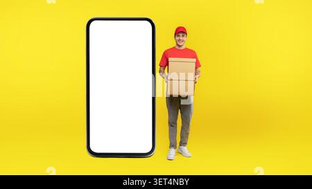 Smiling Young Man Holding Cardboard Box, Pointing At Empty Smartphone Screen, Business Concept Stock Photo