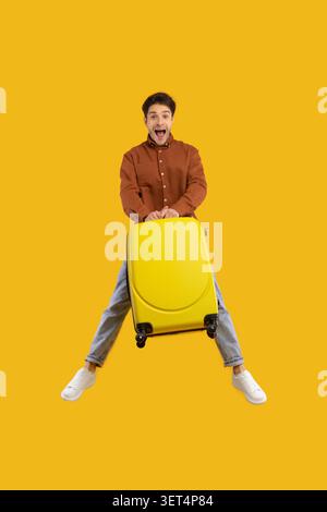 Excited Young Man Holding Yellow Suitcase Jumping, Studio Background Stock Photo