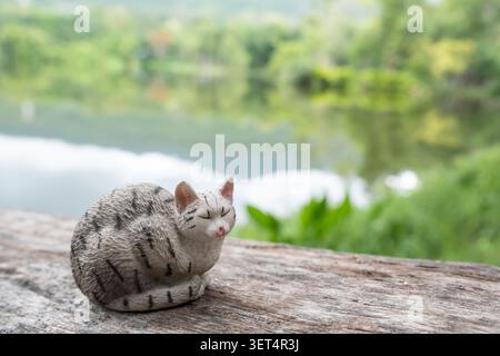 Chiang Mai, Thailand - 19 June 2026 : A grey tabby cat figurine curled up sleeping on a wooden log, with a lake and green forest in the background Stock Photo
