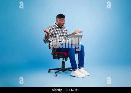 Surprised Black Male Student Using Laptop While Sitting On Chair, Office Setting Stock Photo