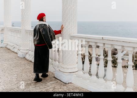 Woman Balcony Sea Stylish woman in a red beret and black jacket stands on a white terrace with lights overlooking the ocean Stock Photo