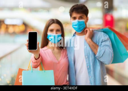 Happy Young Couple Wearing Masks, Holding Smartphone And Shopping Bags In Mall, Unrecognizable Stock Photo