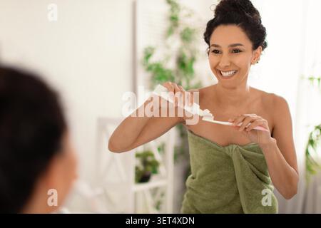 Happy Young Woman Smiling While Brushing Teeth In Bathroom, Home Skincare Routine Stock Photo