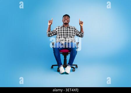 Happy Black Male Sitting In Office Chair, Pointing Up, Excited Expression, Business Concept Stock Photo