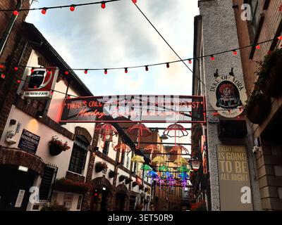 "Neon sign reading 'There's only seven types of rain in Belfast' hangs above a narrow alleyway adorned with colourful umbrellas at the Duke of York." Stock Photo