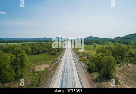 Wide empty road stretches through green landscape in sunny weather near mountains in the background Stock Photo