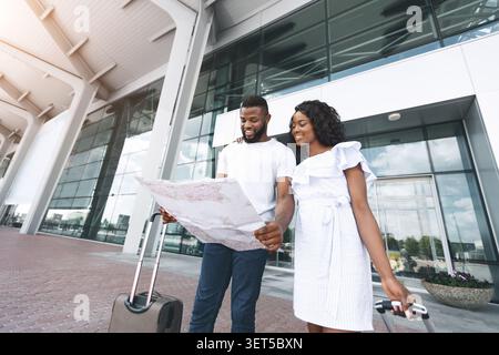 Couple Explores Travel Plans While Standing Outside a Modern Building With a Suitcase Stock Photo