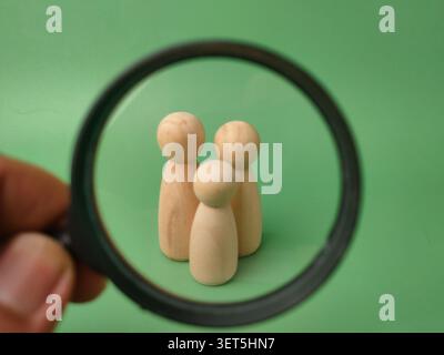A hand holds a magnifying glass over three wooden peg figures standing on a green background. Stock Photo
