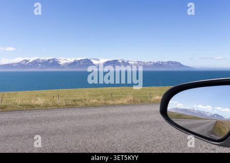 Coastal road along a fjord with snow-capped mountains, reflected in a car wing mirror, North Iceland. Stock Photo