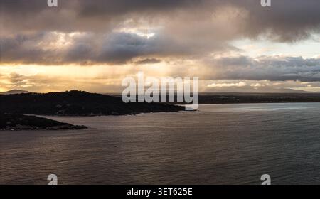 Aerial view of the rugged coastline of Torndirrup National Park under dramatic storm clouds and golden sunset light in Torndirrup, Western Australia, Stock Photo