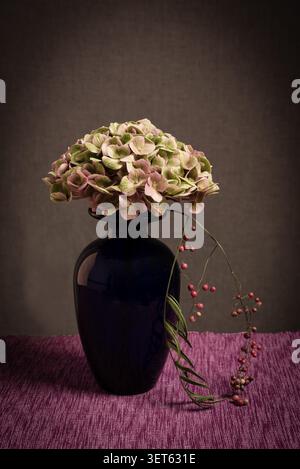Moody fine art still life of a dried hydrangea bloom and red berries in a dark glass vase. Low key lighting. Stock Photo