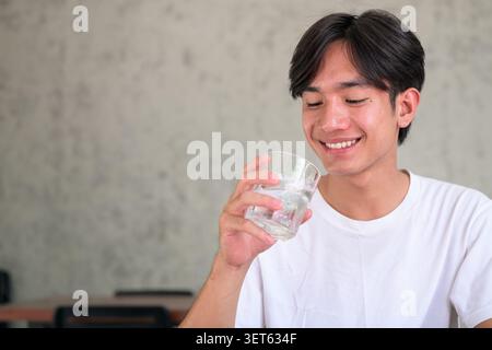 Horizontal lifestyle photograph of a handsome young Southeast Asian Thai Isan non-binary man smiling happily while bringing a glass of water to his mo Stock Photo