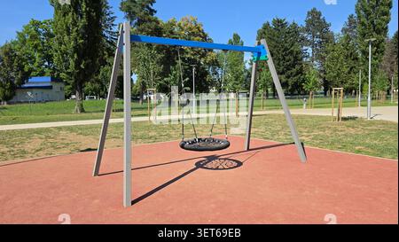 An empty webbed basket swing hangs from metal chains on a grey and blue metal A-frame structure in a sunny municipal park surrounded by grass and tree Stock Photo