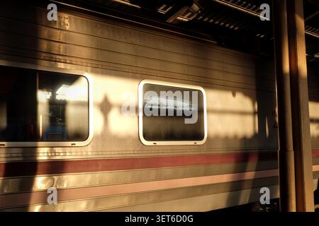 A minimalist close-up of a train passenger car’s metallic side panels and two glass windows, illuminated by dramatic golden hour sunlight and shadows Stock Photo