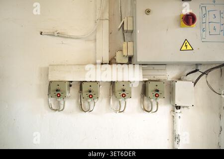 Industrial electrical installation on a white wall. Four grey control boxes with red indicator lights and switches are mounted below a larger grey ele Stock Photo