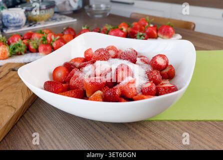 Fresh sliced strawberries covered in granulated white sugar inside large ceramic bowl. Sweet homemade food preparation on wooden kitchen table Stock Photo