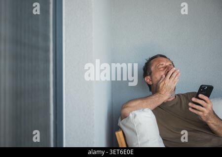 Tired middle-aged man sits on a balcony sofa, yawning and covering his mouth while checking his smartphone, conveying fatigue, boredom, late-night scr Stock Photo