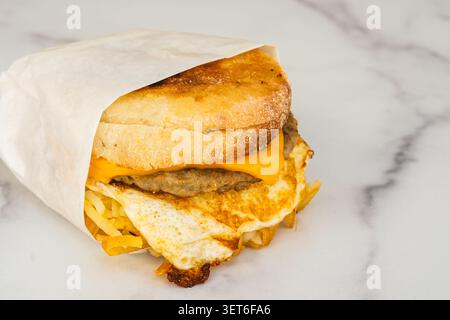 Hot breakfast sandwich with sausage, fried egg, cheddar cheese, and hash browns on a toasted English muffin wrapped in paper. Stock Photo