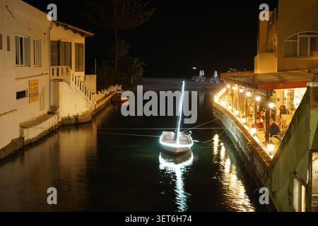 A small fishing boat decorated with neon lights floats at night in a narrow river canal next to a brightly lit waterfront restaurant. Stock Photo