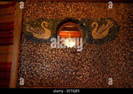 A unique textured mosaic pebble wall featuring two beautiful swans flanking a round window that reflects a glowing crystal chandelier inside. Stock Photo