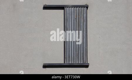A minimalist straight on view of a rectangular window opening on a grey plaster wall partially covered by weathered vertical timber louvers creating Stock Photo