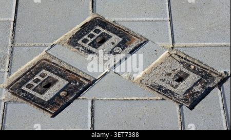 A high angle close up shot of a trio of square metal utility access plates with textured patterns set into a pavement of rectangular concrete paving Stock Photo
