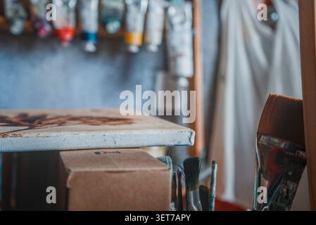 A canvas sits in an art studio next to paint brushes with tubes of paint in the background Stock Photo