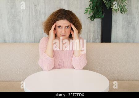 Middle-aged woman holding her head in pain from headache and stress while sitting at a table Stock Photo