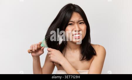 Concerned Asian Woman Brushing Hair, Showing Frustration With Tangles, Home Setting, Hair Care Concept Stock Photo