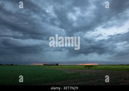 Dramatic dark storm clouds sweep over a flat rural farmland as a thin band of sunset light glows on the distant horizon Stock Photo