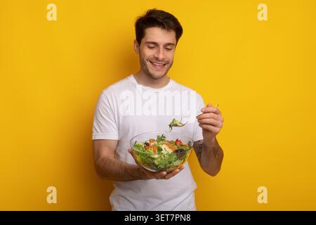 Happy Young Caucasian Man Eating Salad In Front Of Yellow Background, Healthy Eating Concept Stock Photo