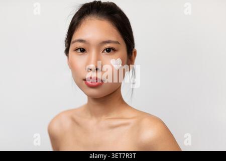 Young Woman Applying Skincare Cream With Heart Shape On Face, Beauty Concept Stock Photo