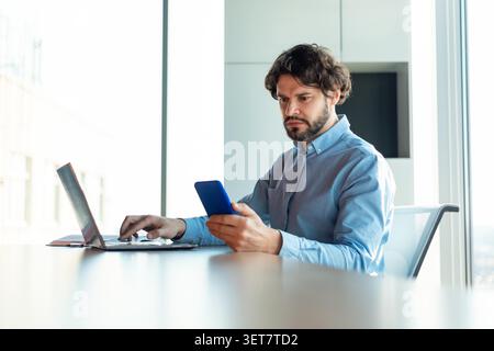 Focused Hispanic Man Using Smartphone While Working On Laptop At Office Desk, Business Concept Stock Photo