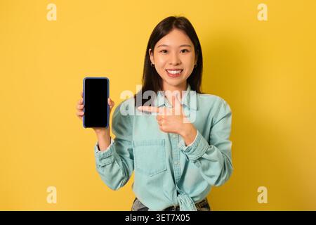 Smiling Asian Woman Pointing At Smartphone With Blank Screen, Yellow Background, Mockup Stock Photo