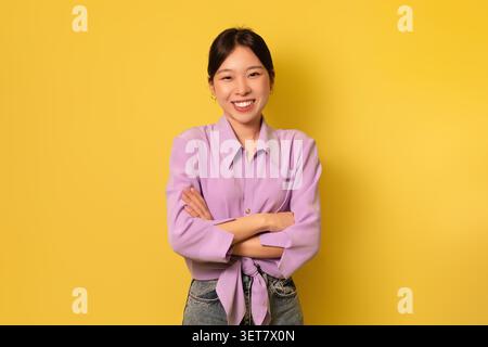 Smiling Young Asian Woman With Arms Crossed, Happy Expression, Yellow Background, Business Concept Stock Photo