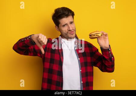 Disappointed Young Caucasian Man Giving Thumbs Down With Burger, Unhealthy Eating Concept Stock Photo