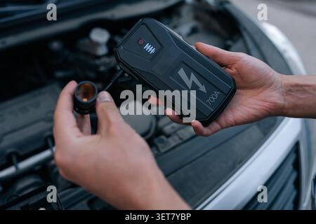 Close-up of hands holding portable car jump starter. Device ready for use with open engine bay in background. Concept for roadside assistance, vehicle Stock Photo